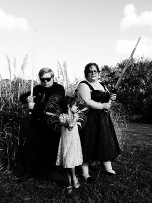 Black and white image of a family of three in wedding regalia. The parents hold lightsabers and stand back to back while the child holds a blaster pistol and aims towards the camera in front of them.